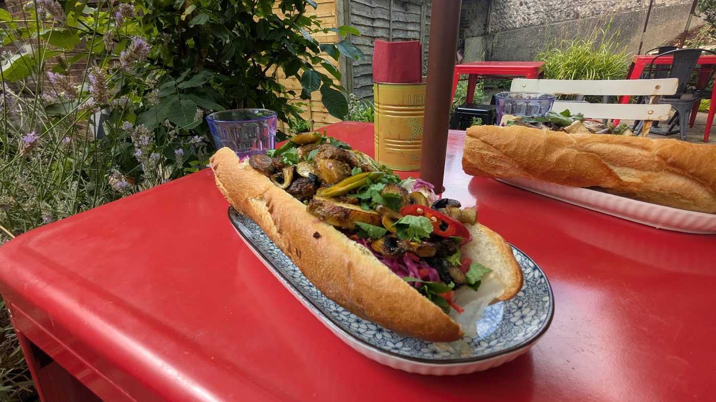 Two bánh mì on a red table outside the shop, with greenery and the street behind.