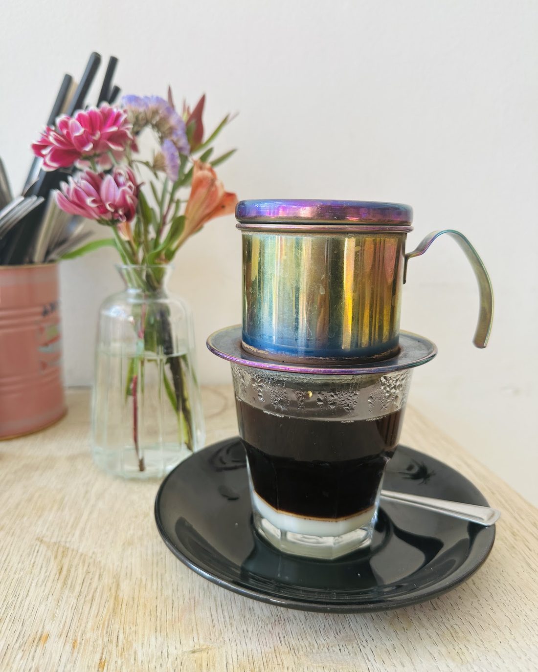 A Vietnamese phin filter dripping coffee into a glass on a saucer, beside a jar of flowers.